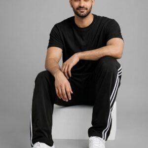 "A man wearing a black t-shirt and black track pants with three white side stripes sits sideways on a white block. He is wearing white athletic shoes and looking ahead against a plain light gray studio background."